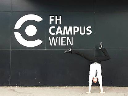 Mann in weißen Hemd, Krawatte und Anzugshose vor einer Wand, auf der das F H Campus Wien Logo angebracht ist, welches aus dem künstlerisch gezeichnete C-Form und daneben der Schriftzug Hochschule Campus Wien besteht. Der Mann macht gerade einen Handstand und formt dabei mit seinen Füssen ein Häkchen.