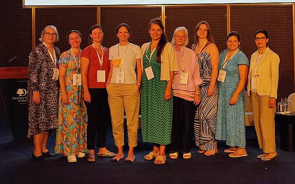 © privat Neun Personen stehen gemeinsam auf einer Bühne bei einer Konferenz, alle tragen Namensschilder und sommerliche Kleidung