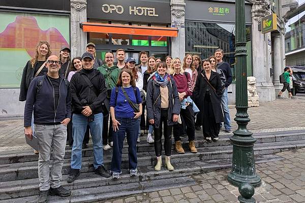 © Martina Behringer Gruppenfoto einer Exkursionsgruppe im städtischen Raum: Rund 25 Personen stehen gemeinsam auf den Stufen vor einem Gebäude und blicken in die Kamera. Im Hintergrund sind Geschäftslokale und die Fassade eines Hotels zu sehen. Die Teilnehmenden tragen Freizeitkleidung und wirken entspannt. Die Aufnahme entstand bei bewölktem Wetter in einer europäischen Innenstadt.