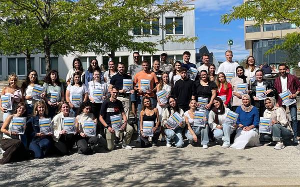 © Hochschule Campus Wien Eine große Gruppe junger Menschen posiert auf Stiegen, sie halten bunte Mappen in der Hand. Im Hintergrund ist ein Gebäude sowie Himmel zu sehen.