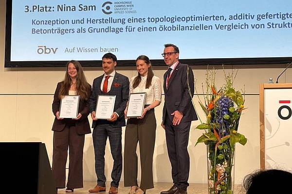 © HCW/Link Vier Personen stehen auf einer Bühne und halten Urkunden; im Hintergrund ist eine Präsentationsfolie mit dem 3. Platz und einem Projekttitel zu sehen, daneben ein Blumenarrangement.