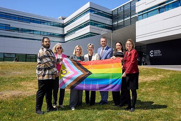 © HCW/Schedl Sechs Personen stehen vor dem Gebäude der Hochschule Campus Wien auf einer Wiese und halten gemeinsam eine Progress-Pride-Flagge. Im Hintergrund ist die moderne Hochschulfassade bei sonnigem Wetter zu sehen. Die Gruppe blickt in die Kamera und setzt ein sichtbares Zeichen für Vielfalt und Inklusion.